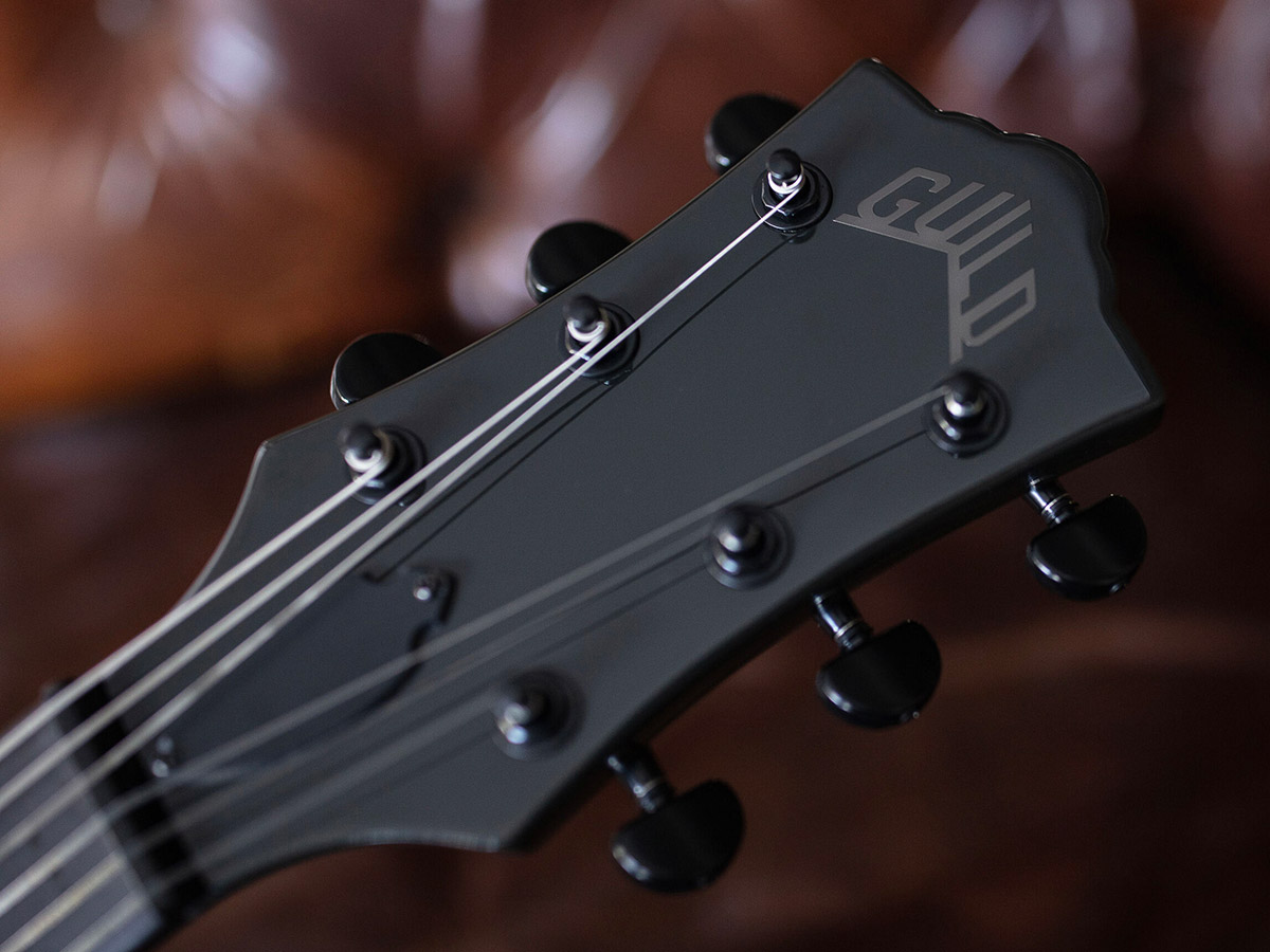 Close-up of Polara Night Edition Tungsten guitar headstock with Guild Peak logo in front of a leather couch