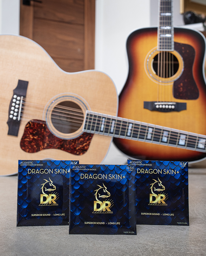 Three Dragon Skin+ String packages in front of two Guild USA acoustic guitars