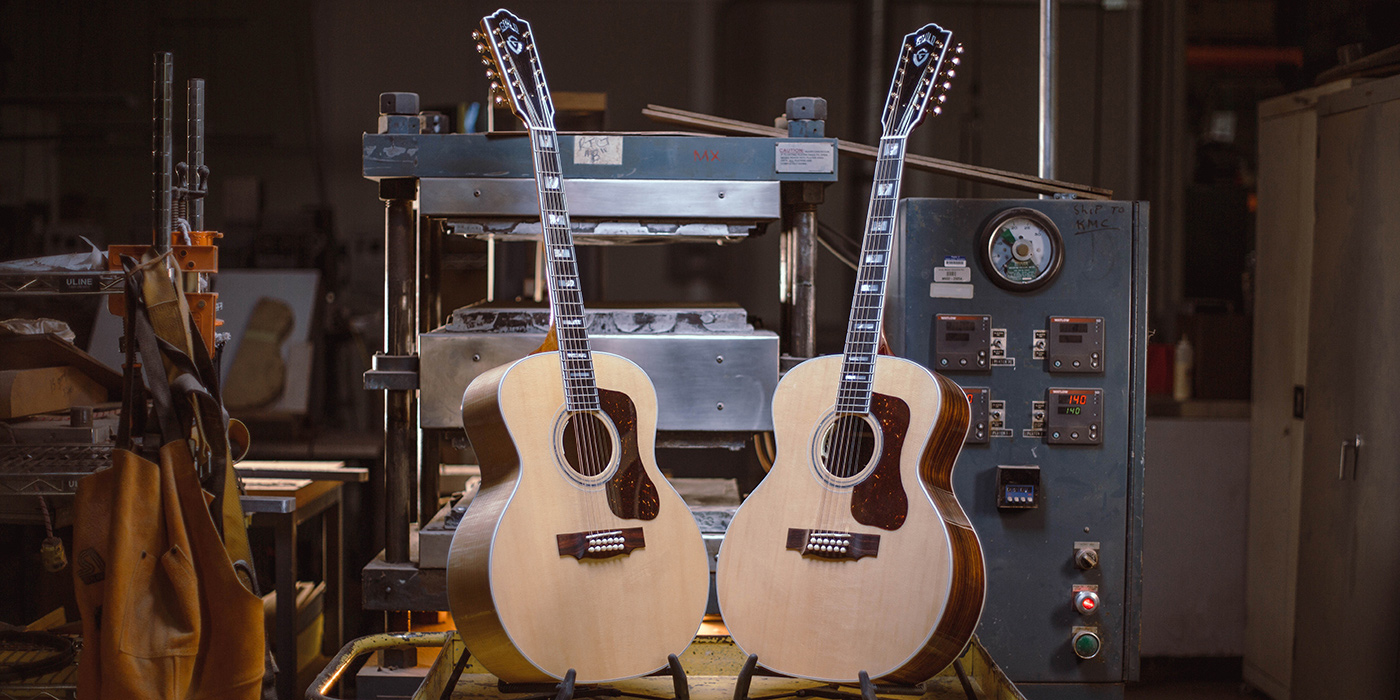F-512 acoustic guitars are displayed in guitar workshop, with a machinery backdrop and tools nearby. The setting is well-lit and industrial.