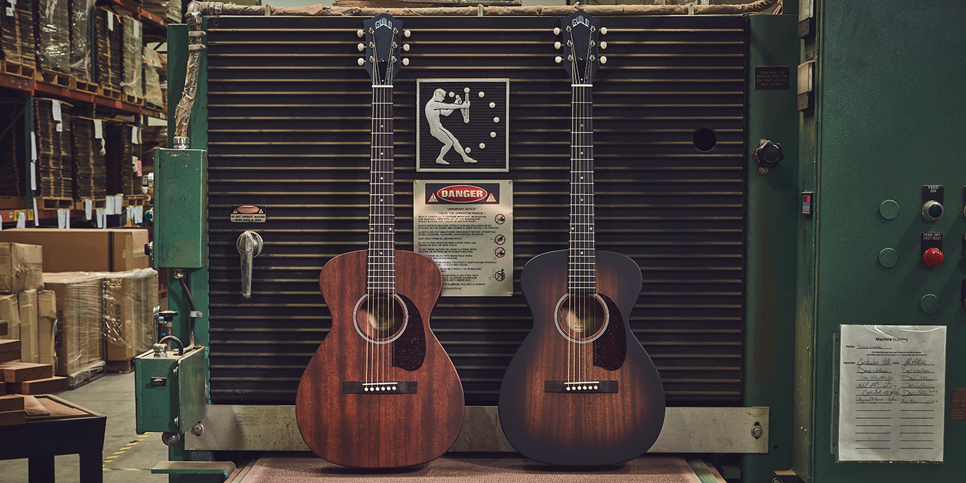 M-20 Natural and M-20 Vintage Sunburst acoustic guitars are displayed in guitar workshop, with machinery and safety signs in the background.
